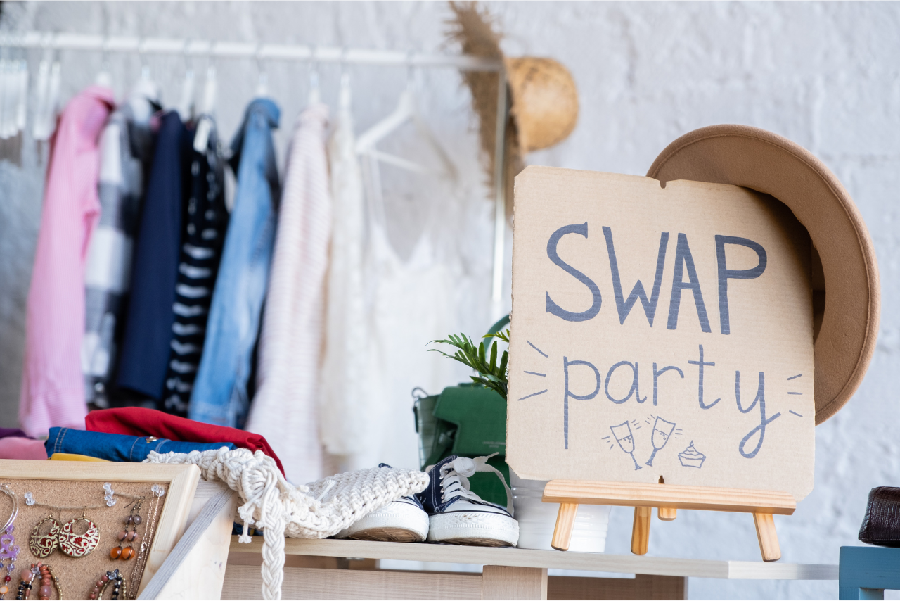 A rack of sweaters in the background with a sign in front that says Swap Party. A hat is hanging from the corner of the sign.